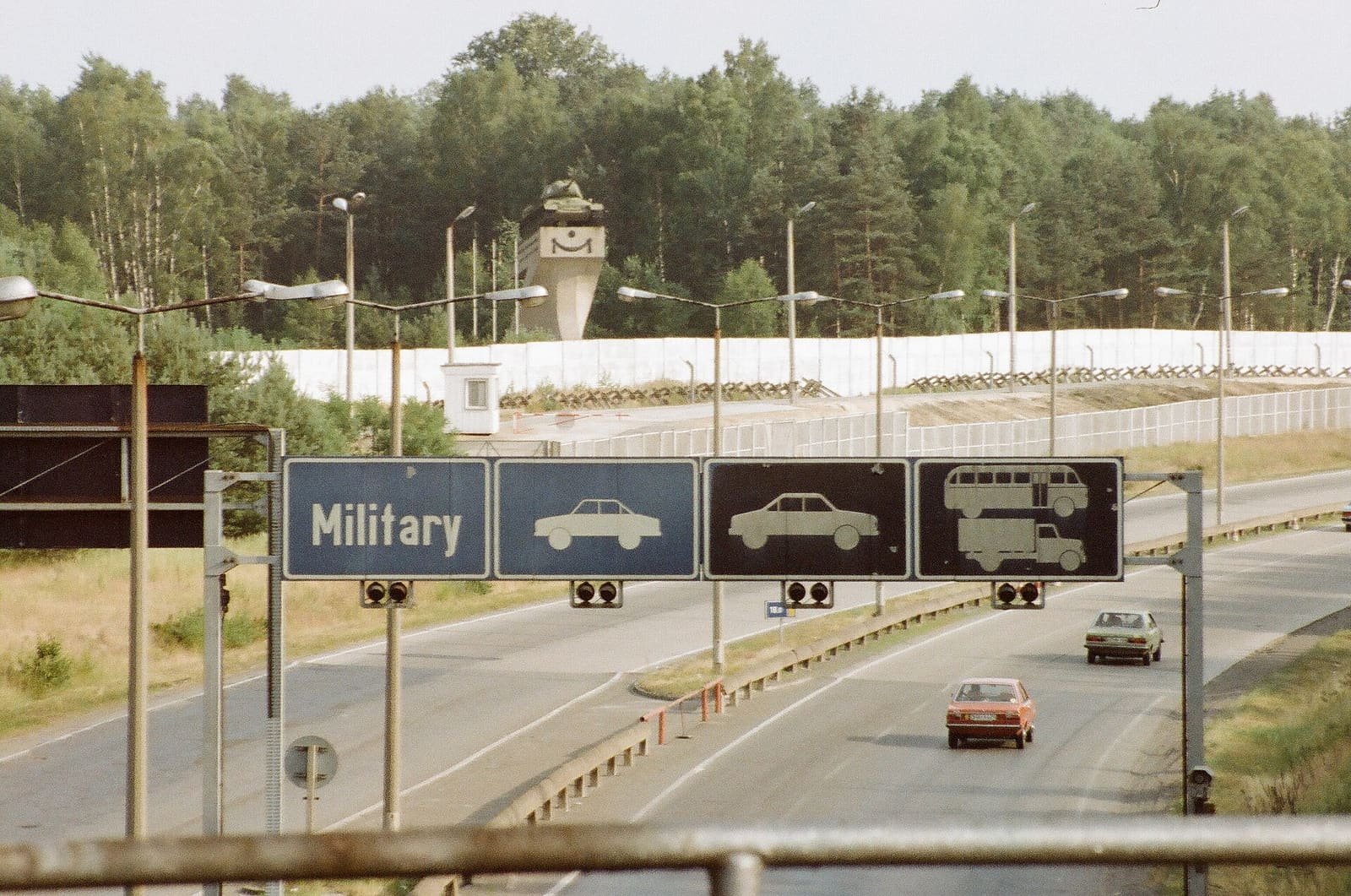 Colour photo of an autobahn with overhead signs reading 'Military' and car, lorry and bus symbols; a white border wall and a tank monument on a plinth are behind.