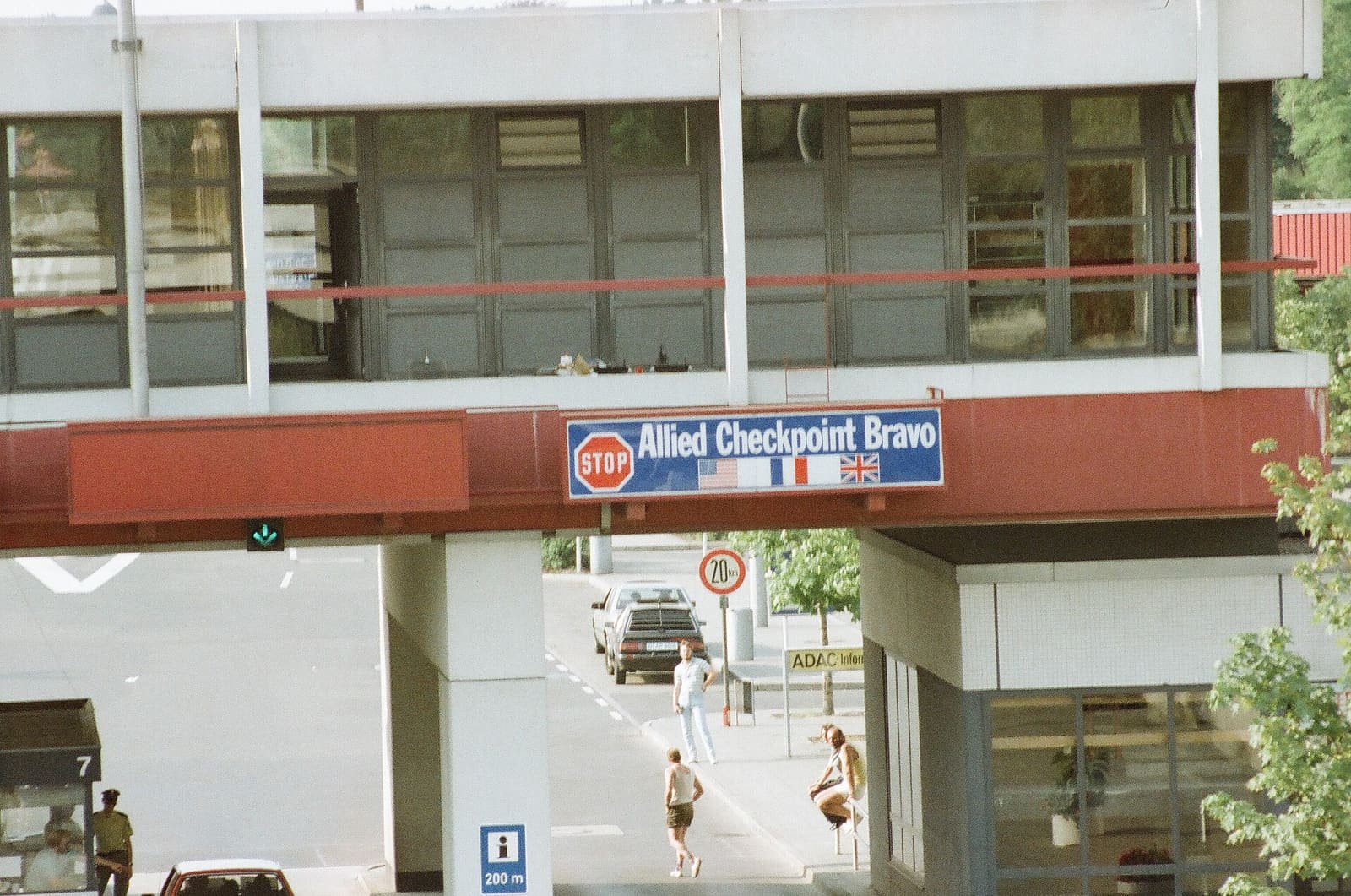 Colour photo of a bridge building over the road with a blue sign reading 'Allied Checkpoint Bravo' and US, French and British flags; people walk beneath it.