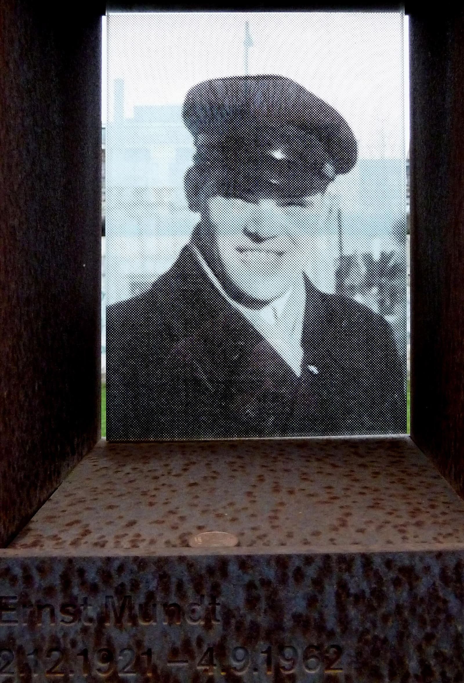 A rust-coloured steel memorial wall with rows of small portrait windows; one shows a black-and-white portrait of Ernst Mundt