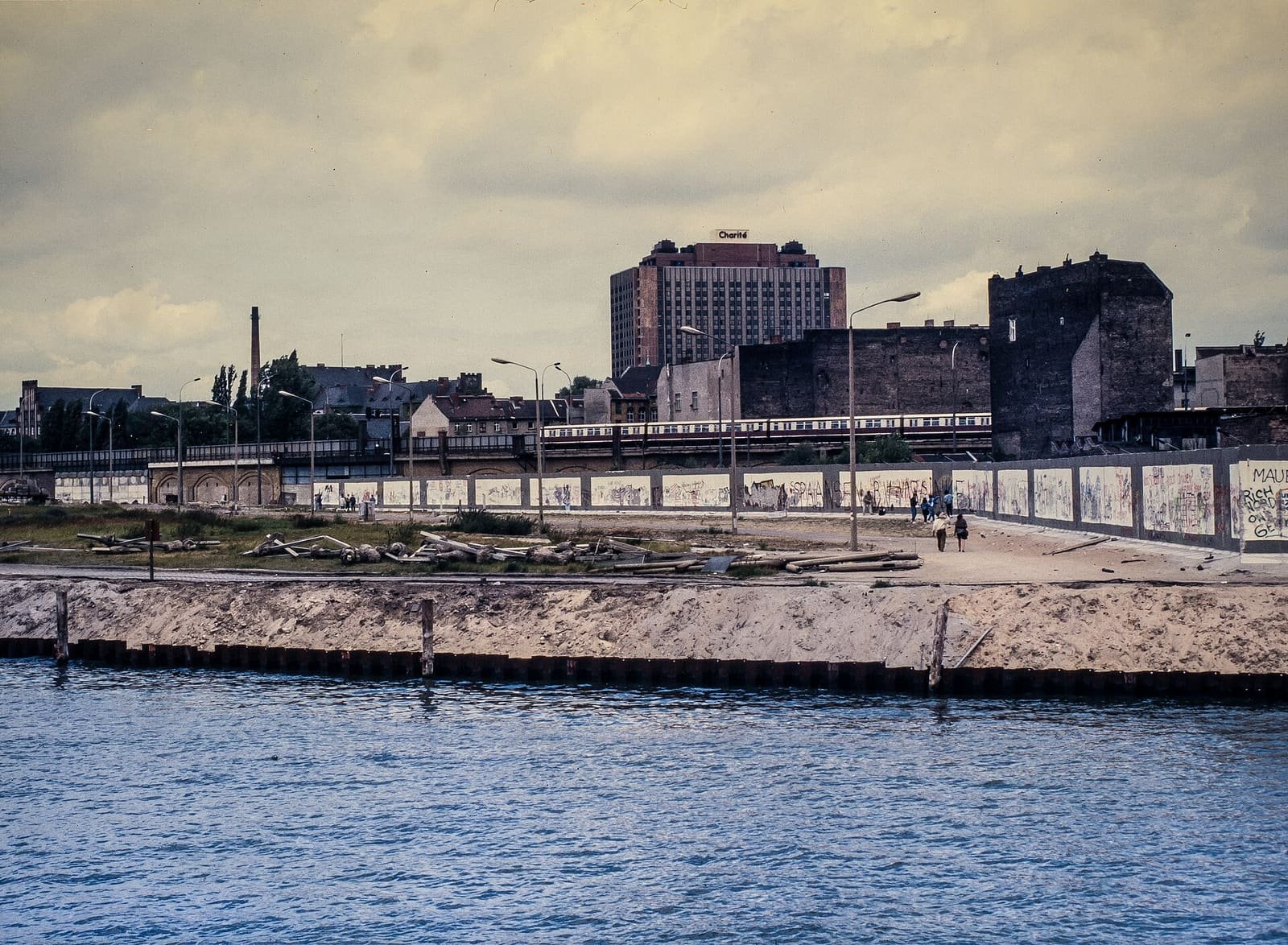 A river in the foreground and, on the opposite bank, a sandy strip and a long graffiti-covered wall, with an elevated railway and a high-rise hospital beyond.