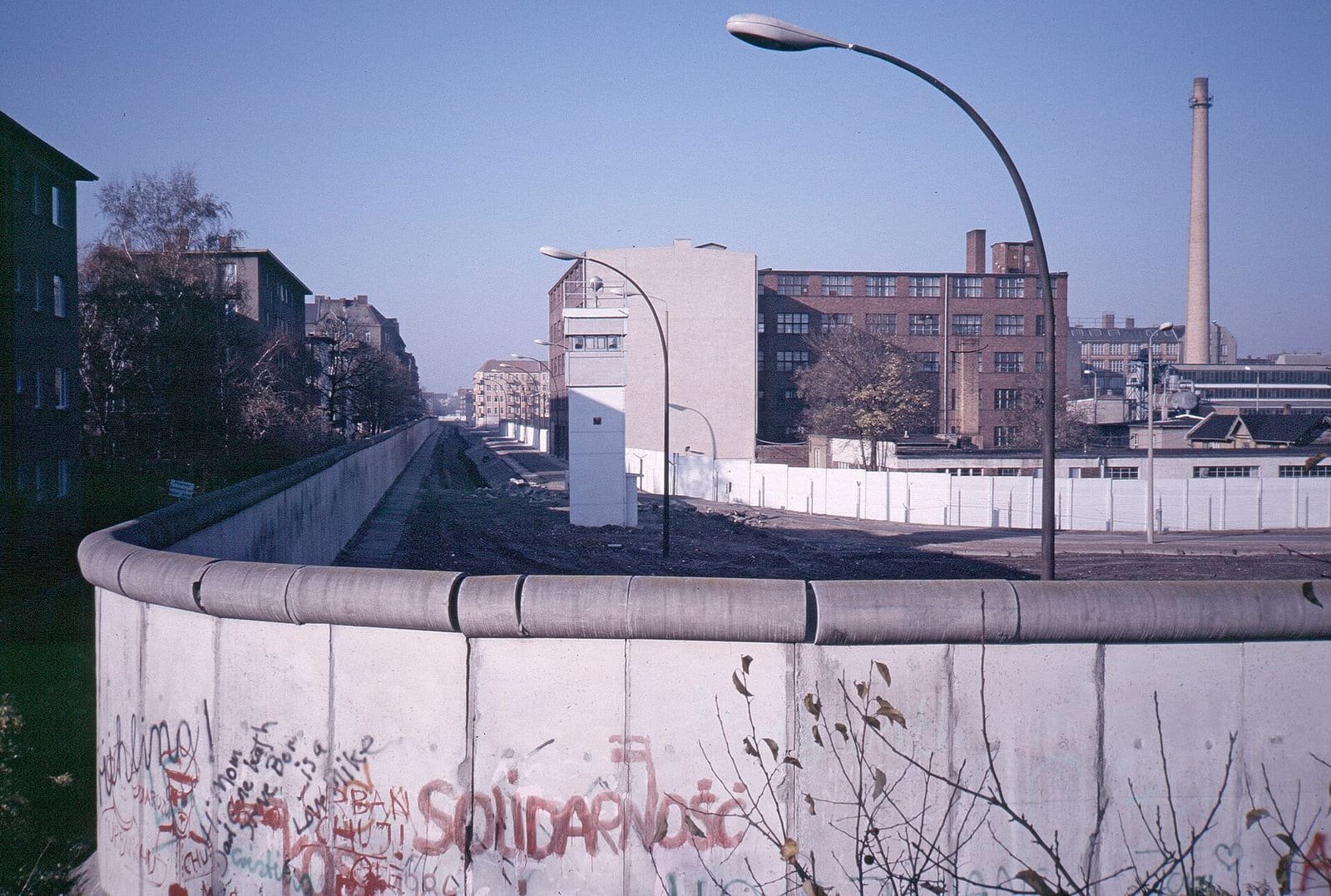 Colour photo: a curved concrete wall with graffiti including 'Solidarność' in the foreground; behind it a cleared strip with a watchtower and lamp posts runs between apartment blocks and factory buildings.