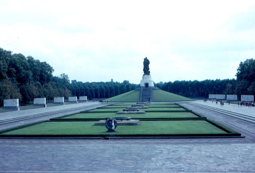 Colour photo of a long formal memorial ground with lawns and stone sarcophagi leading to a grass mound topped by a giant bronze soldier statue, framed by rows of trees.