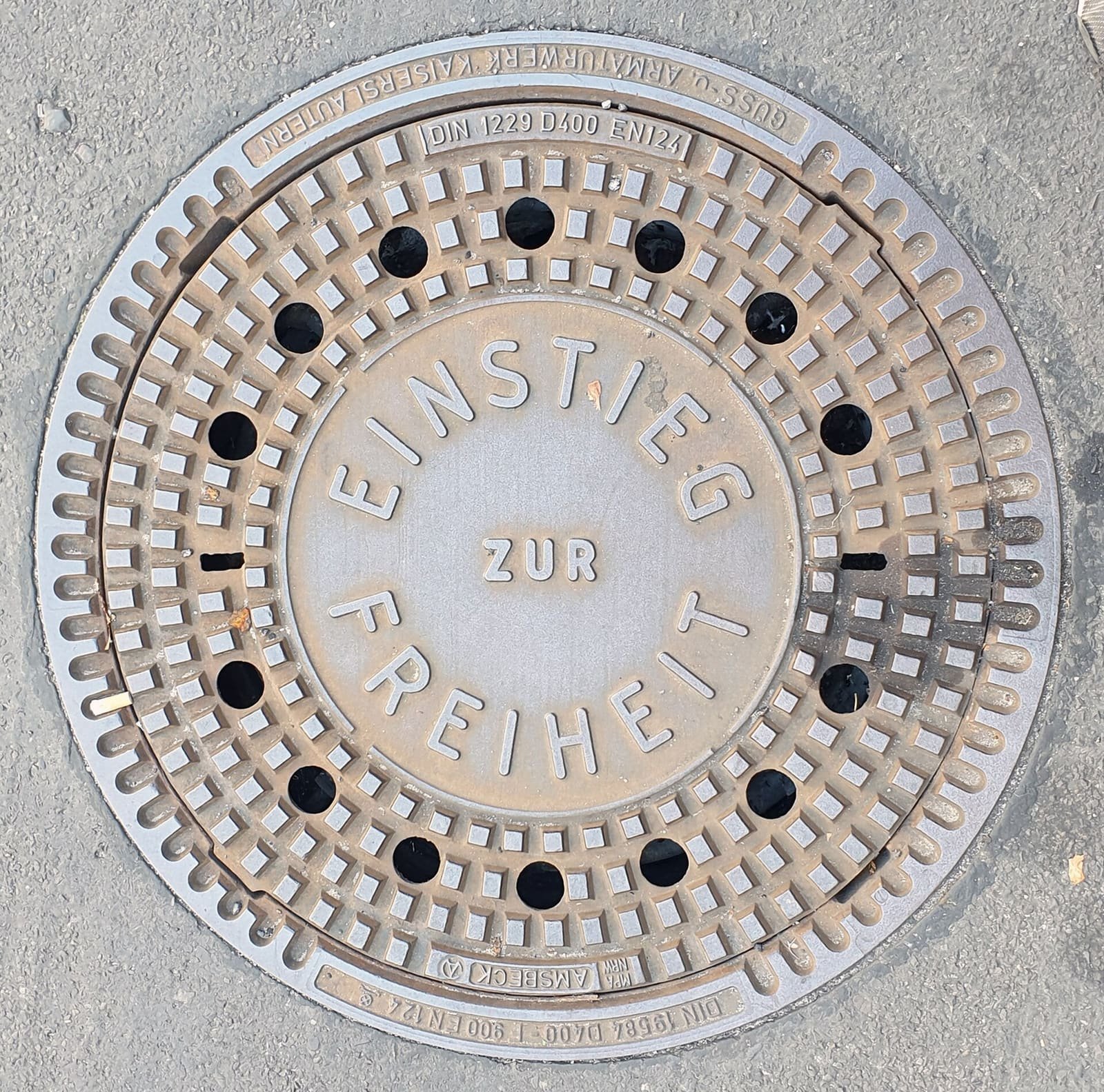 Close-up of a round cast-iron manhole cover set in asphalt, with the raised words 'Einstieg zur Freiheit' in its centre and a ring of holes around the edge.