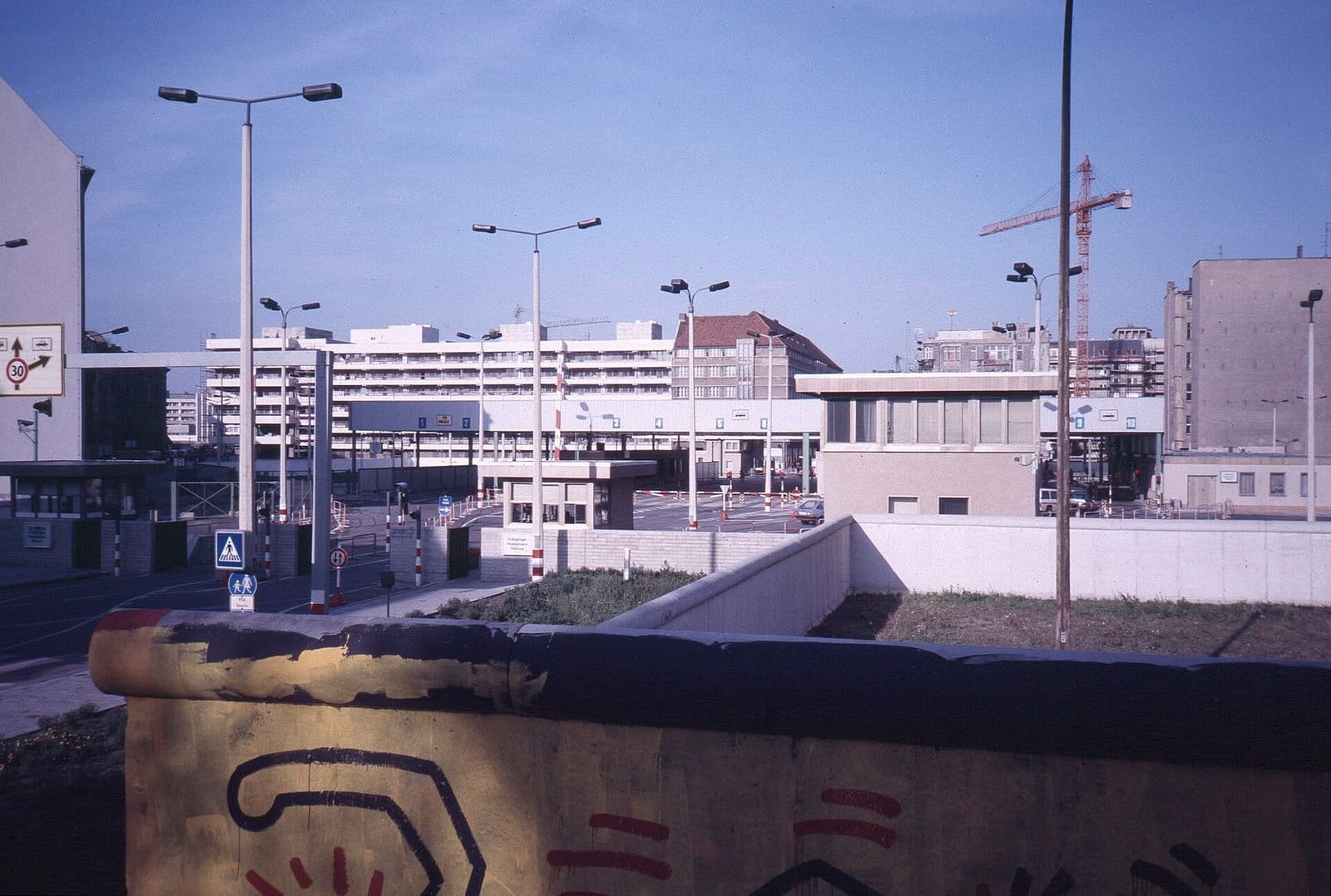 Colour photo over a painted section of the Wall towards the East German control buildings and street lamps at Checkpoint Charlie.