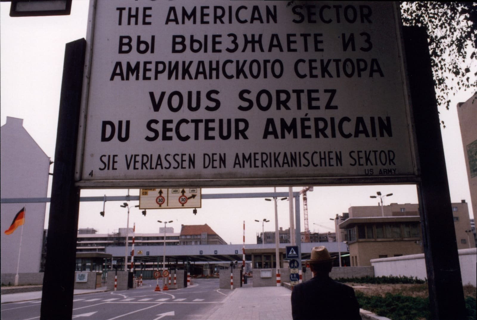 Colour photo of a large four-language sign with the East German checkpoint buildings and barriers visible beneath it.