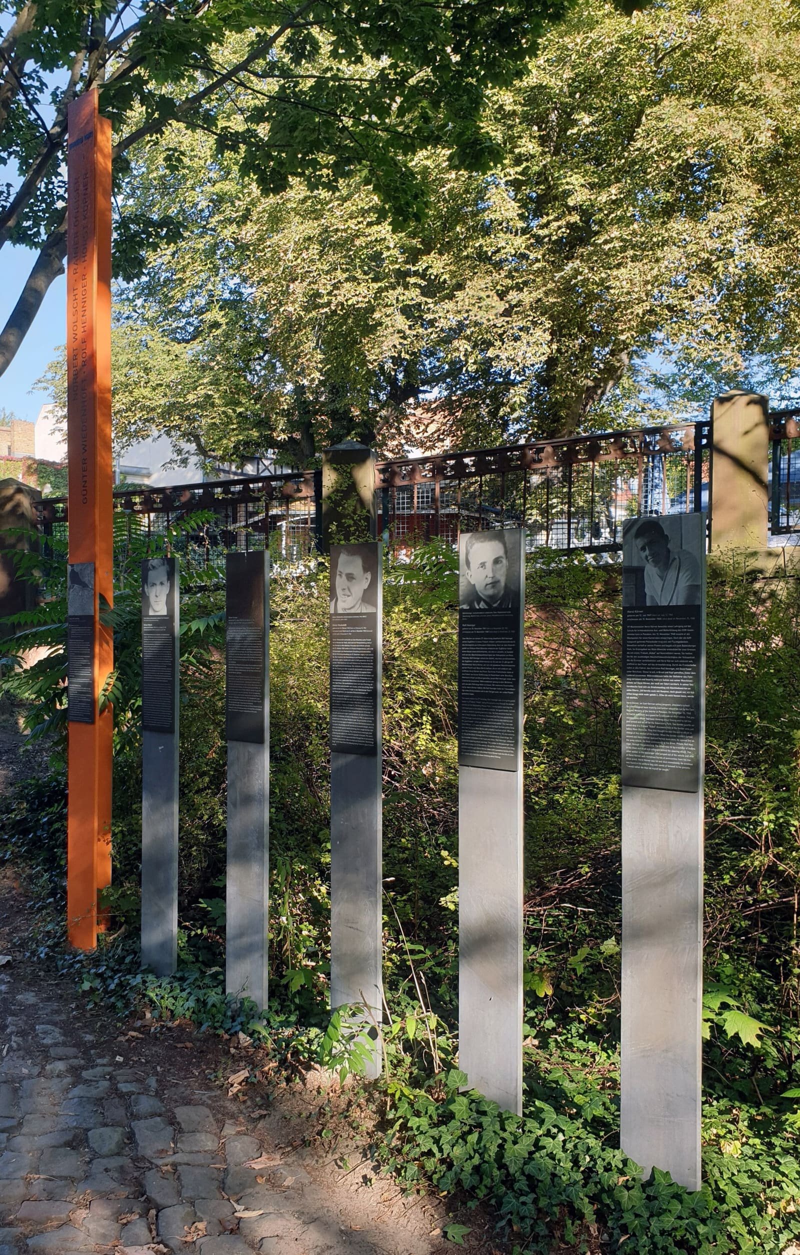 A tall orange stele with names in vertical lettering and five grey steel posts, several bearing portrait photos and text panels, beside a cobbled path under leafy trees.