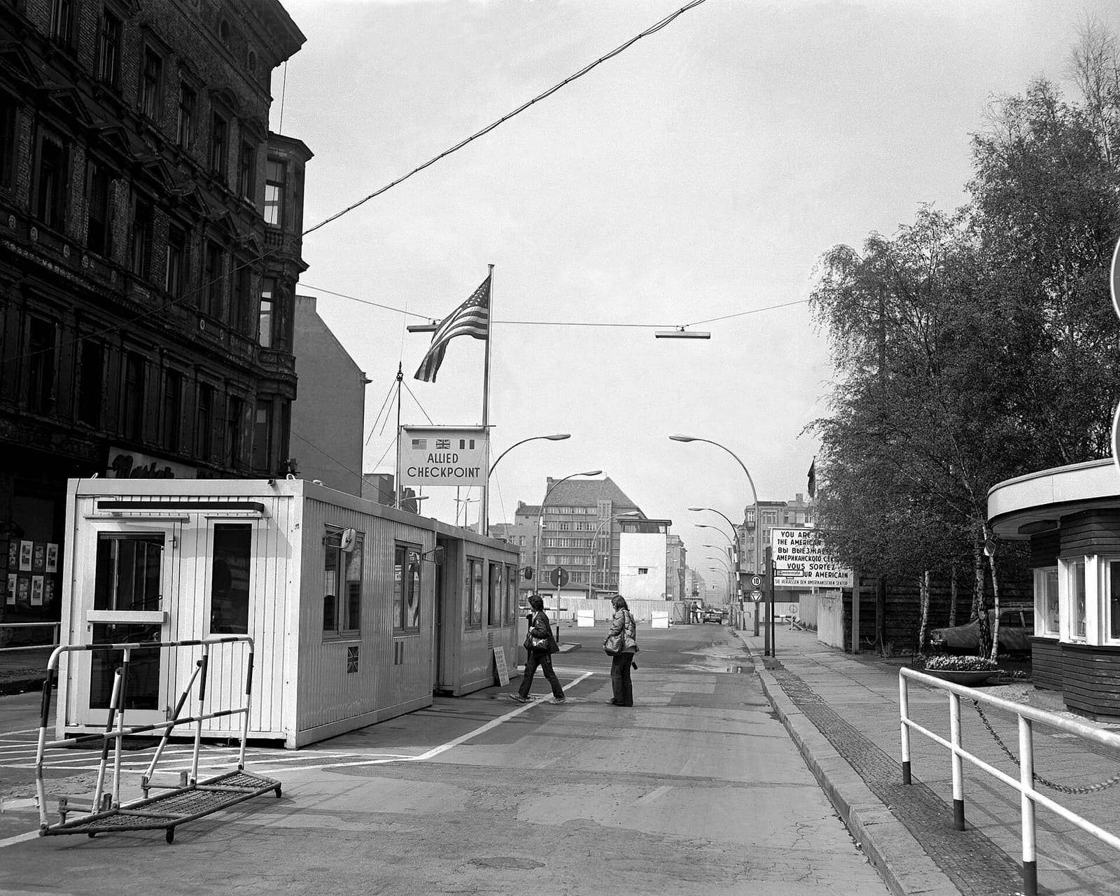 Black-and-white photo of a street with a white prefabricated checkpoint hut under a US flag and an 'Allied Checkpoint' sign, and two pedestrians crossing.