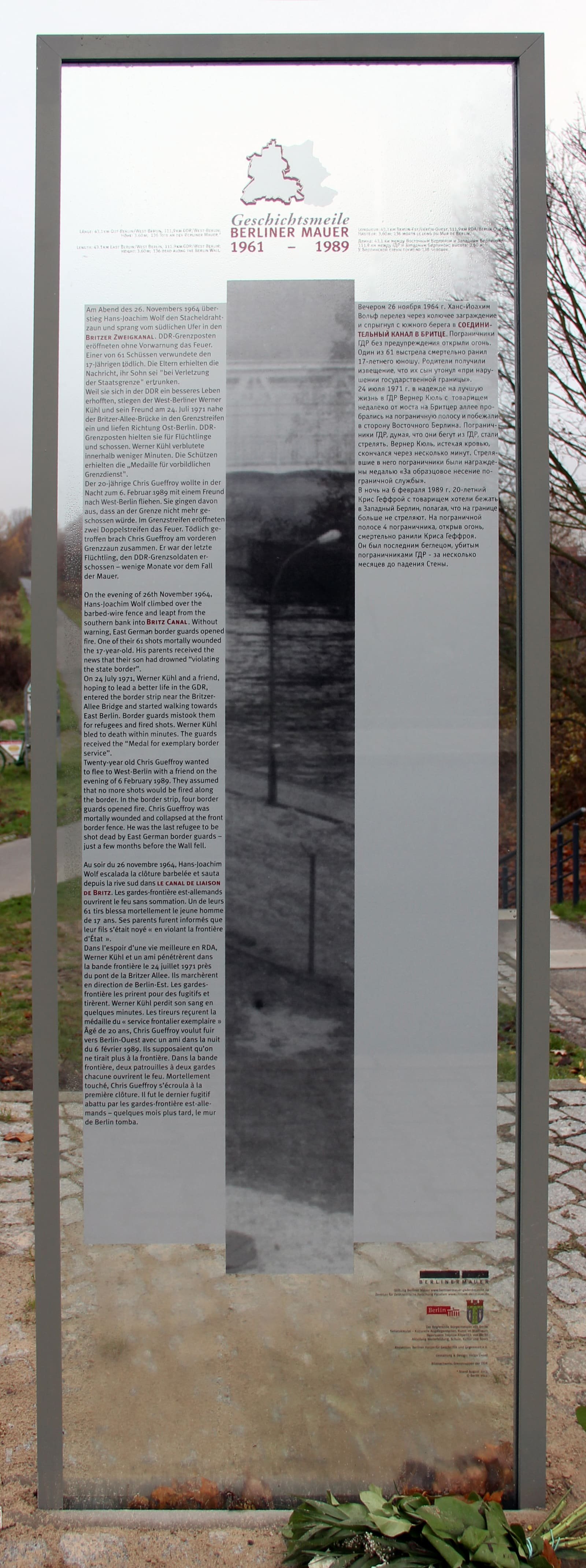 A tall glass panel in a grey frame printed with columns of text in several languages and a faint historical photograph, standing beside a path with bare trees.