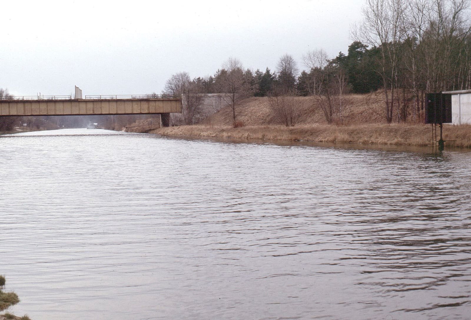 A view along a canal towards a bridge, seen from a footpath on the bank.