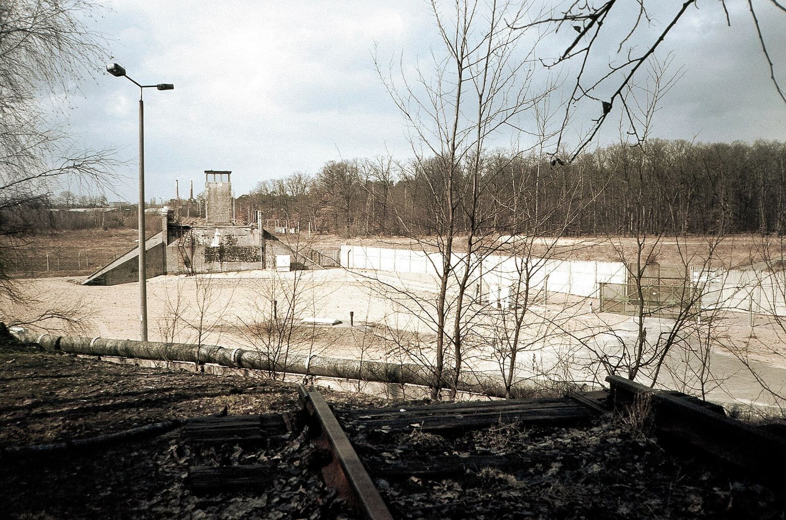 Colour photo from an overgrown railway embankment with an old rail in the foreground: behind a concrete wall lies a wide sandy border strip with lamp posts, a watchtower, a white inner wall and a fence, with woods beyond.