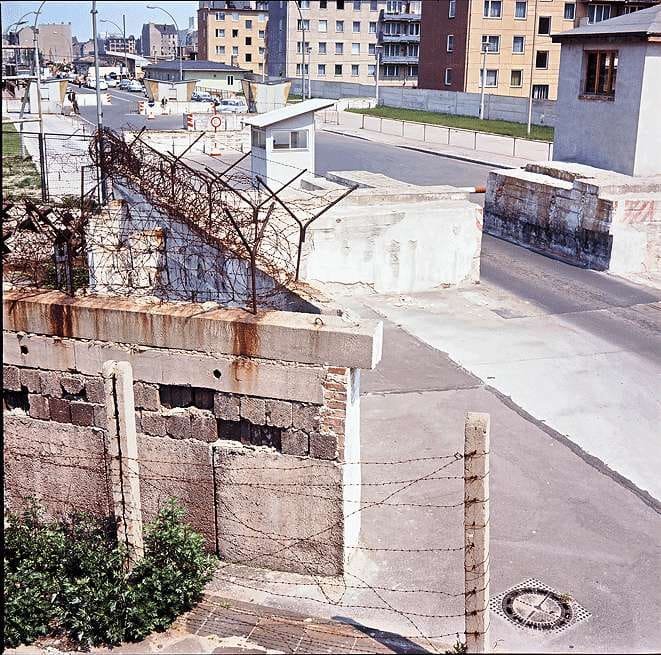 Colour photo taken from above: a wall topped with barbed wire in the foreground, behind it a road with staggered concrete barriers, a small guard hut and a barrier pole, and East Berlin apartment blocks in the background.
