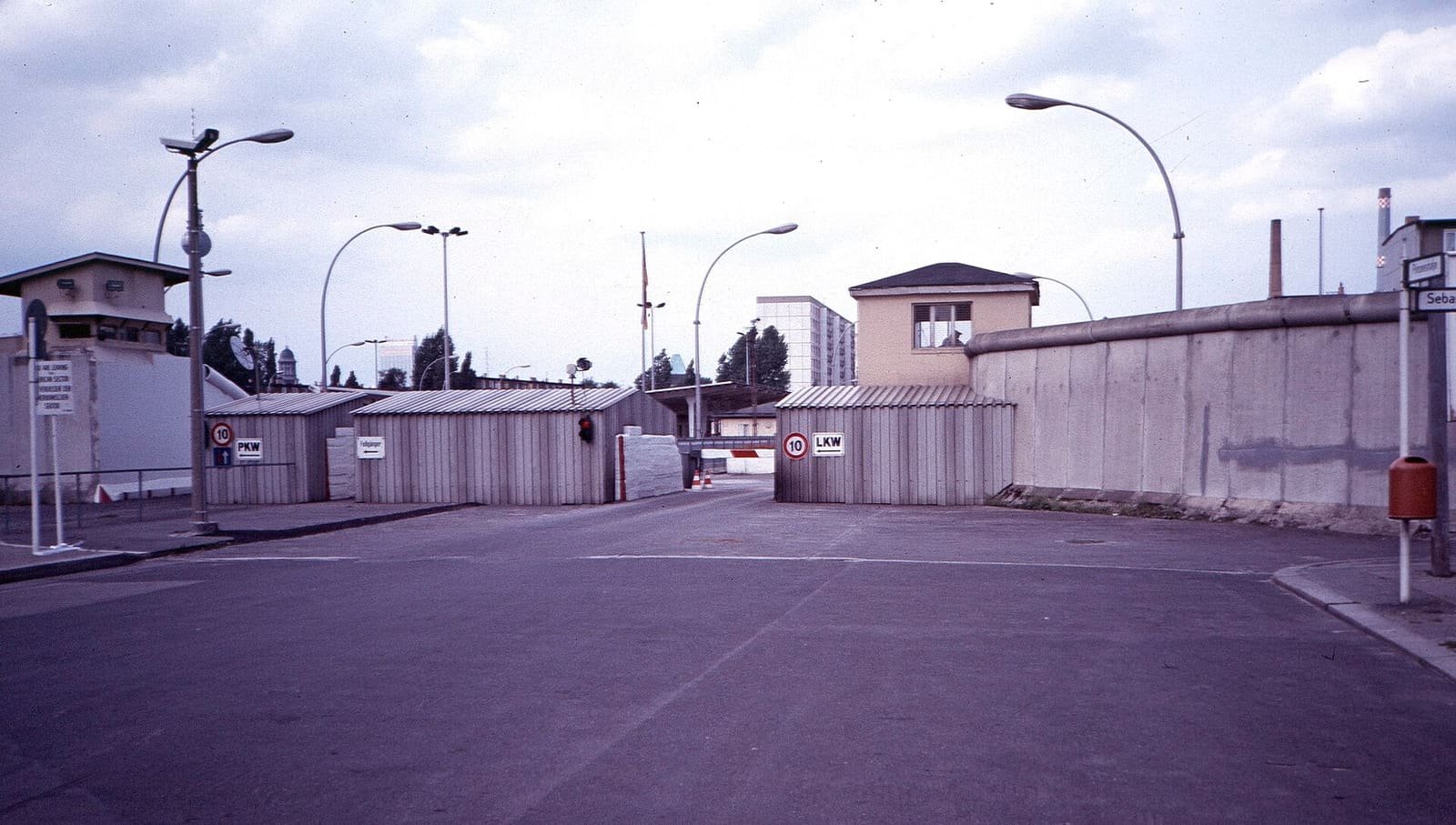 Wide view of a border checkpoint: a road leading between the grey concrete Wall and low control buildings, with a watchtower and street lamps.