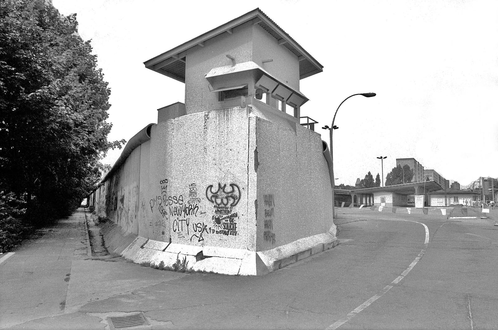 Black-and-white photo of a rounded concrete wall section covered in graffiti with a small control building on top, beside an empty road.