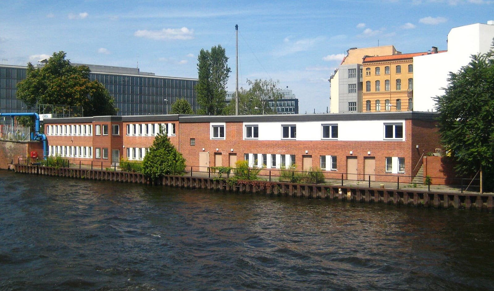 Colour photo: a long low red-brick building with white-framed windows stands on a quay wall at the edge of a river, with office buildings and trees behind it.