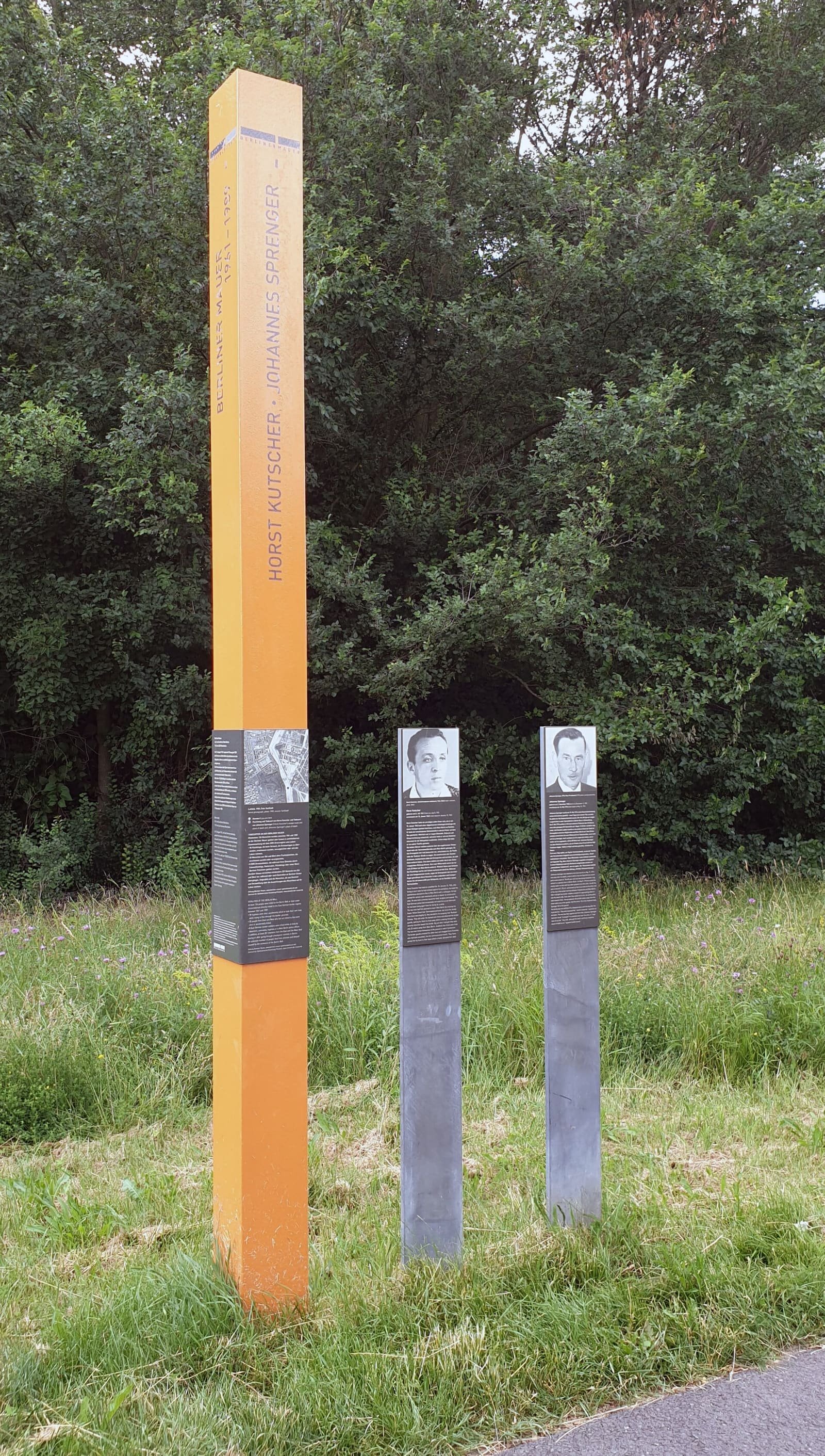 An orange stele and two grey posts with portrait panels standing in grass in front of dense dark-green trees.