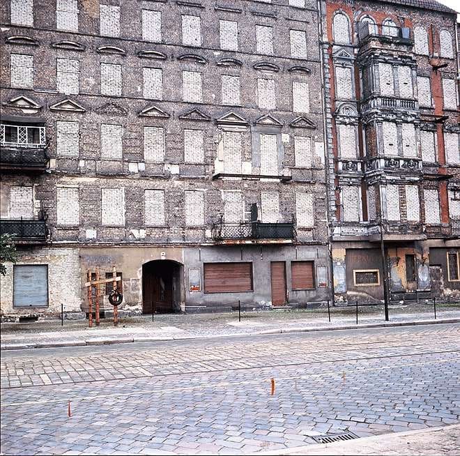 A house front with bricked-up windows, with a memorial on the pavement in front.