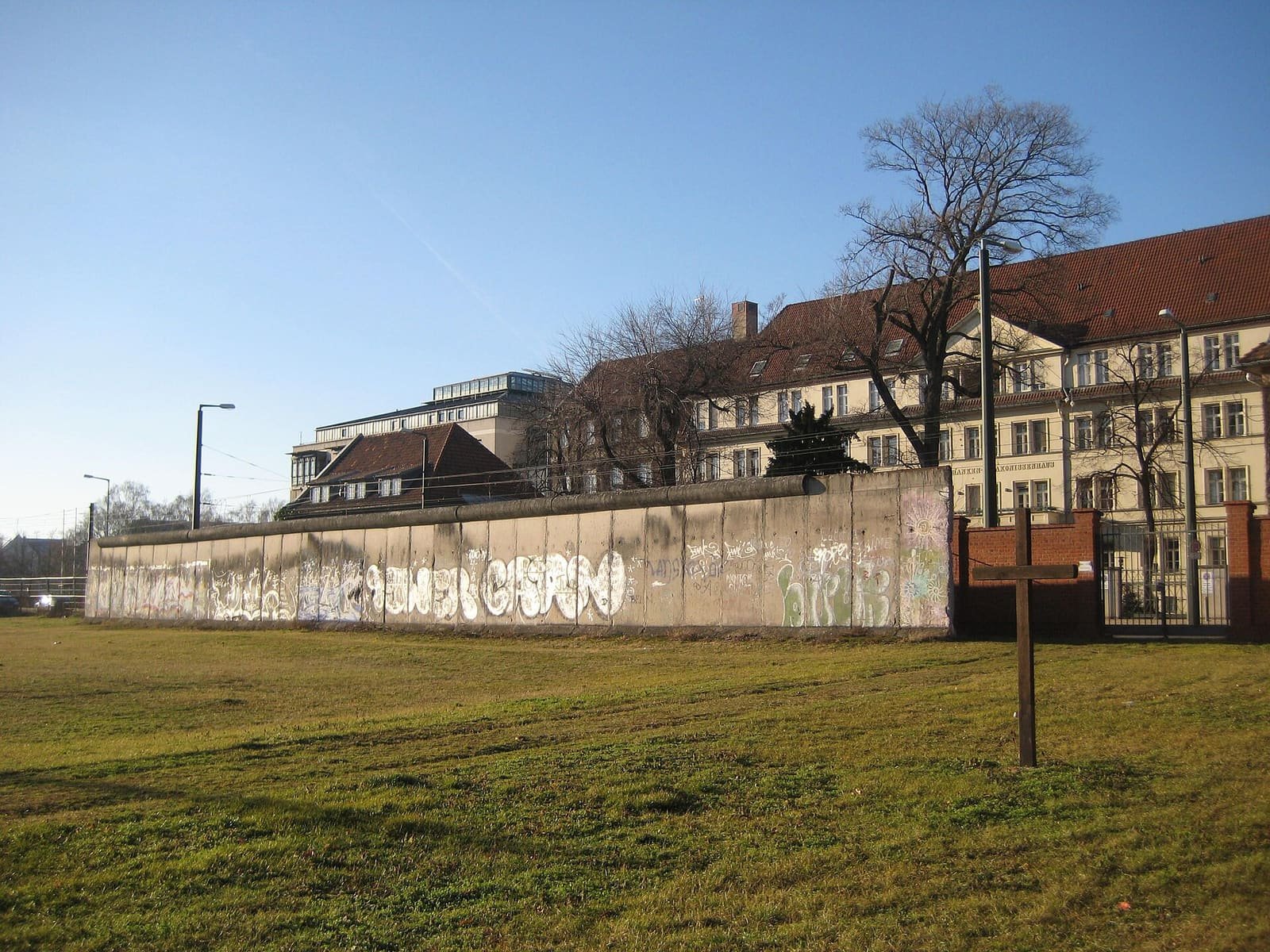 A long grey concrete wall covered in graffiti runs across a grassy field; a simple wooden cross stands in the foreground and large buildings with red-tiled roofs rise behind the wall.