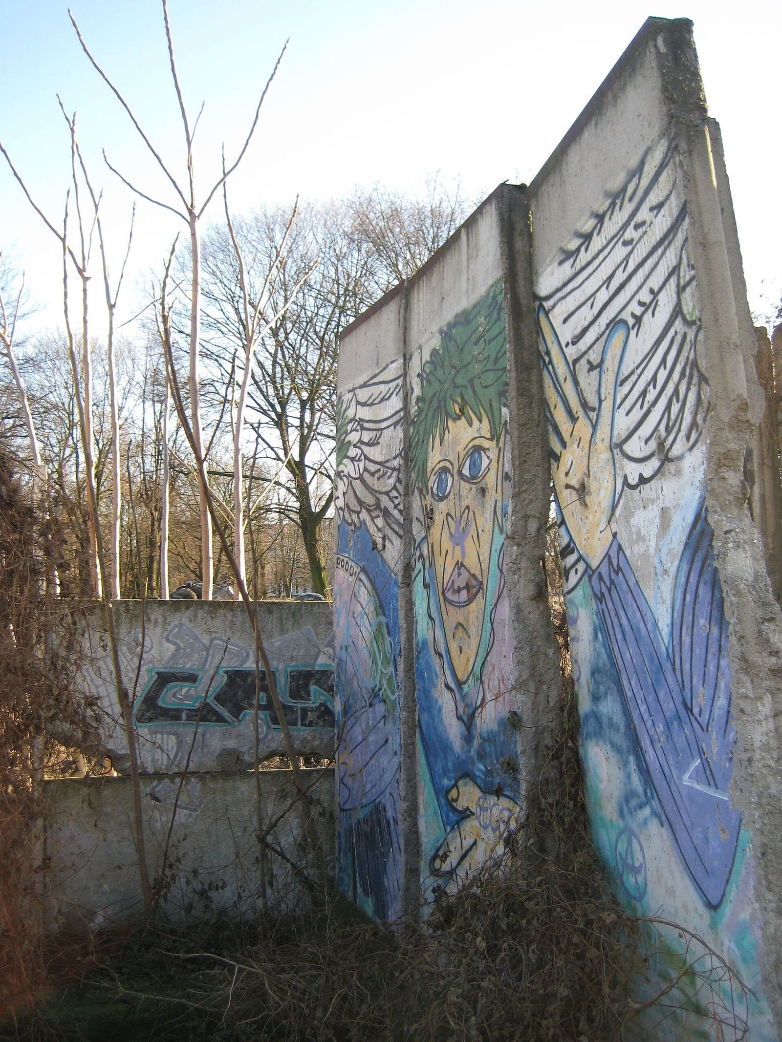 Two tall concrete wall segments painted with a cartoon face and a large hand stand among bare shrubs; behind them on the left runs a lower, grey, graffiti-covered wall.