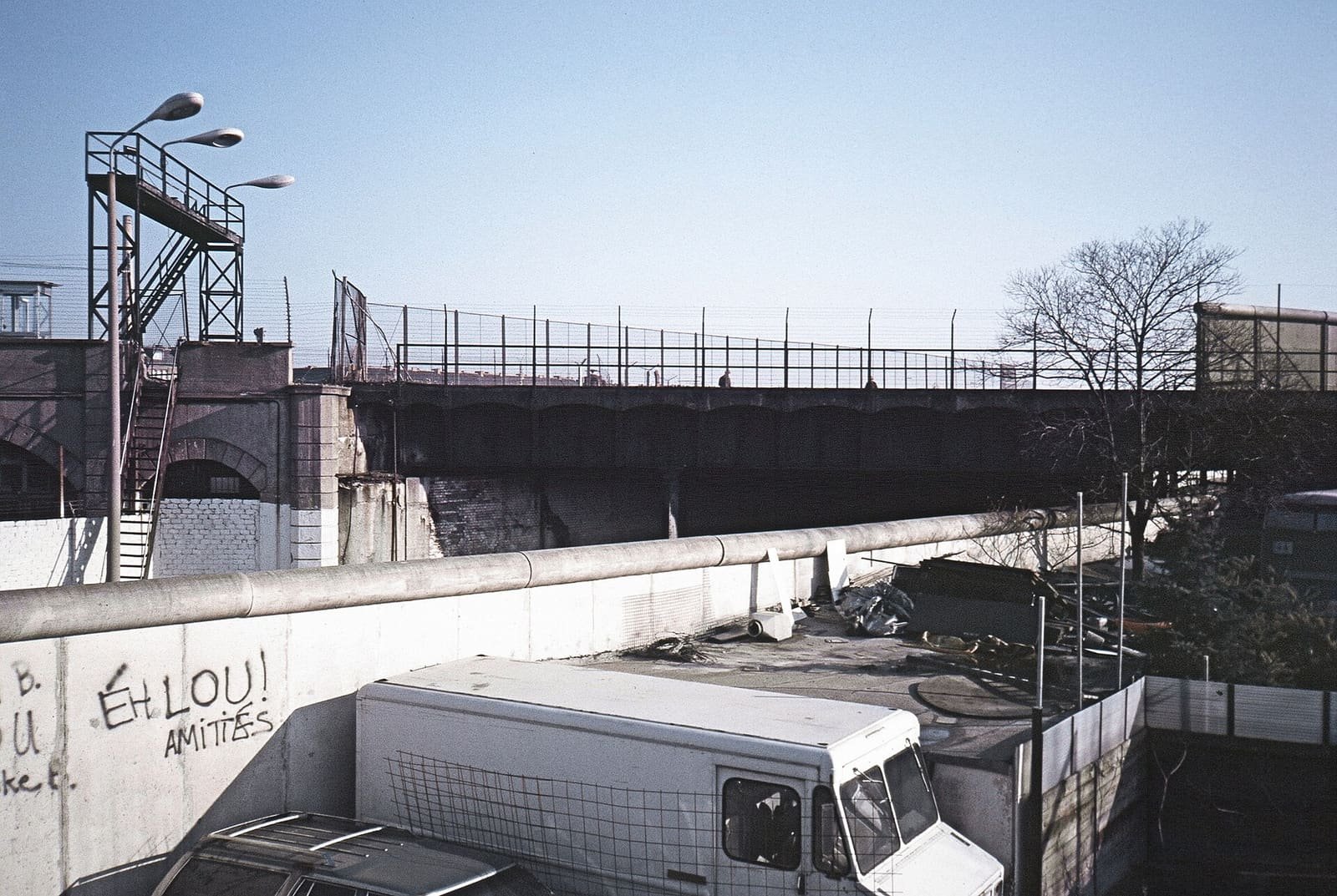 Colour photo over a graffiti-marked concrete wall: a railway bridge with a high fence on top, a lamp platform with stairs on the left and a white van parked below in the border area.