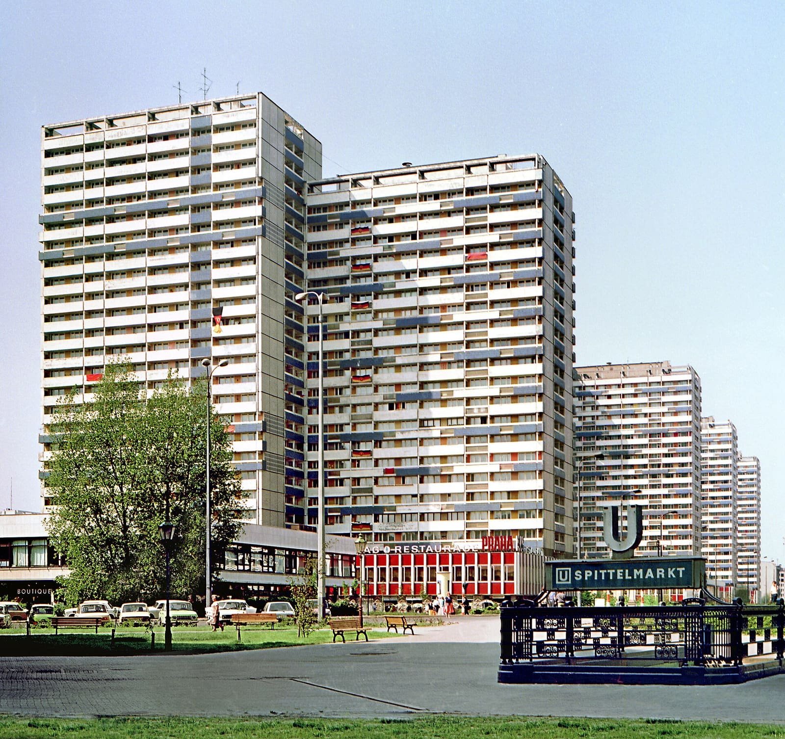 Colour photo: a row of tall white prefabricated tower blocks under a clear sky, with a low restaurant pavilion, parked cars and a 'U Spittelmarkt' underground sign in the foreground.
