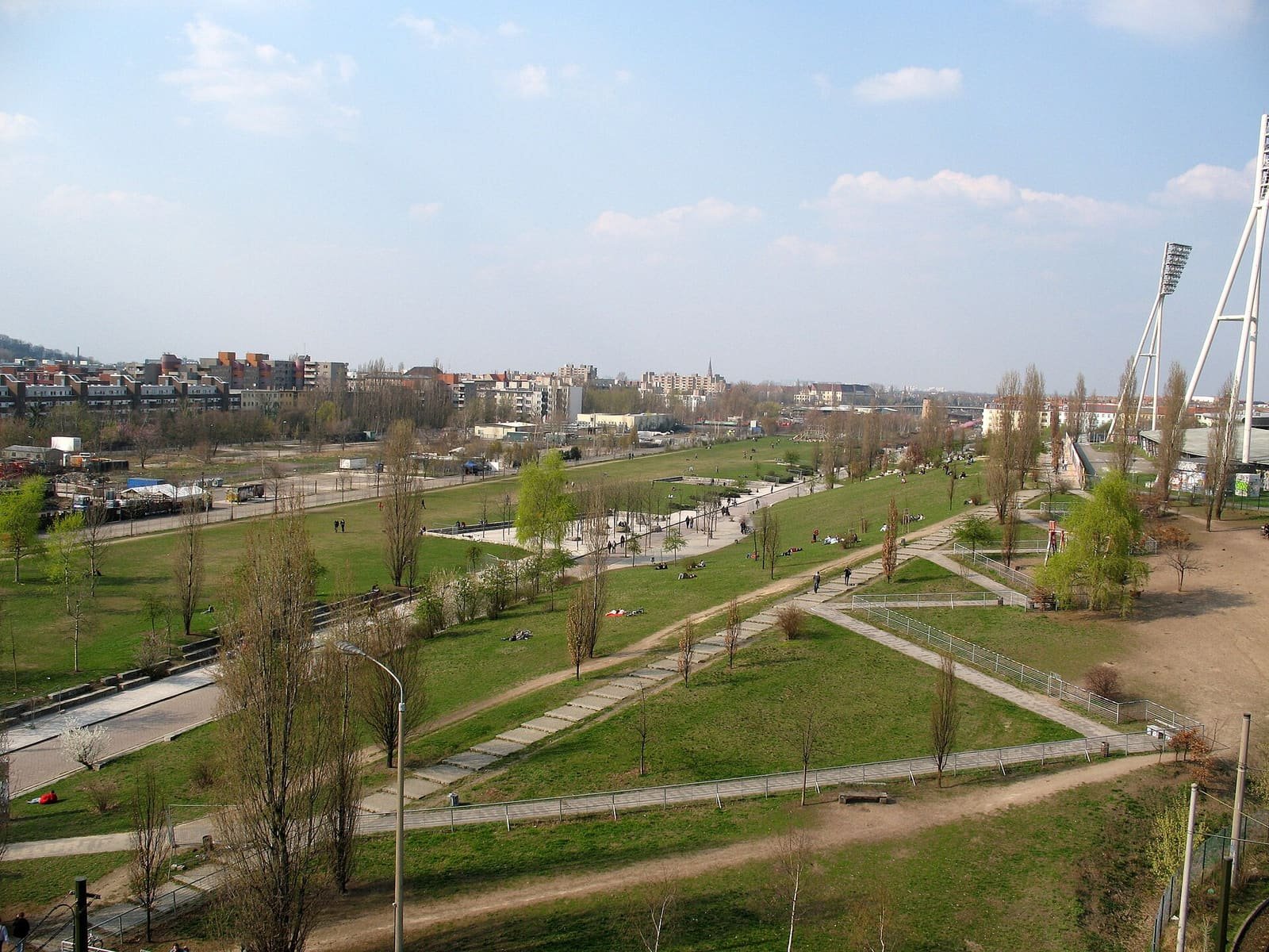 Colour photo from a height: a broad park with lawns, paths and young trees stretches away towards city buildings under a blue sky; two tall floodlight masts rise on the right.