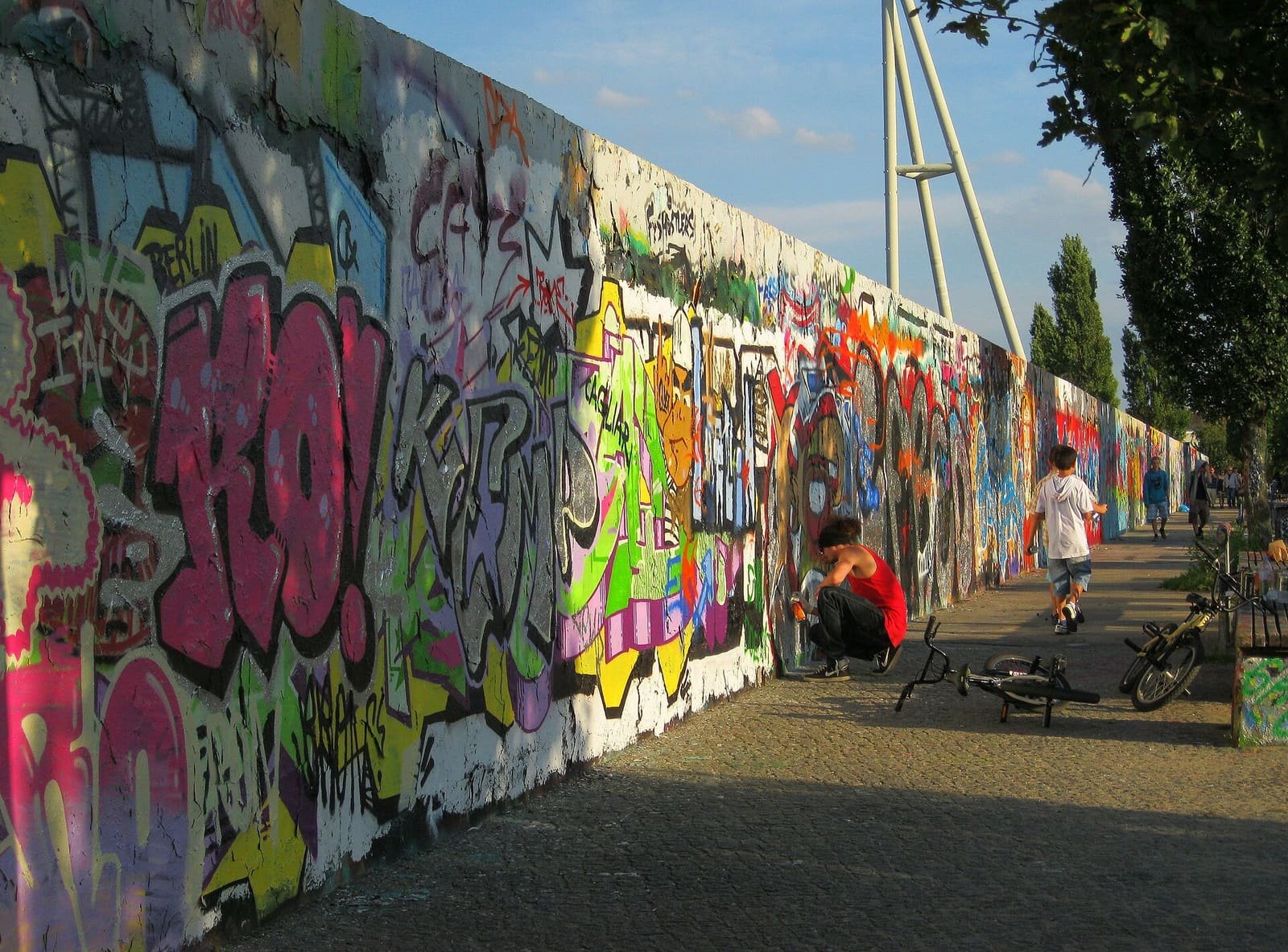 Colour photo: a long concrete wall covered in colourful graffiti runs along a paved path; a person in red crouches at the wall and a boy walks past, with a tall floodlight mast behind the trees.