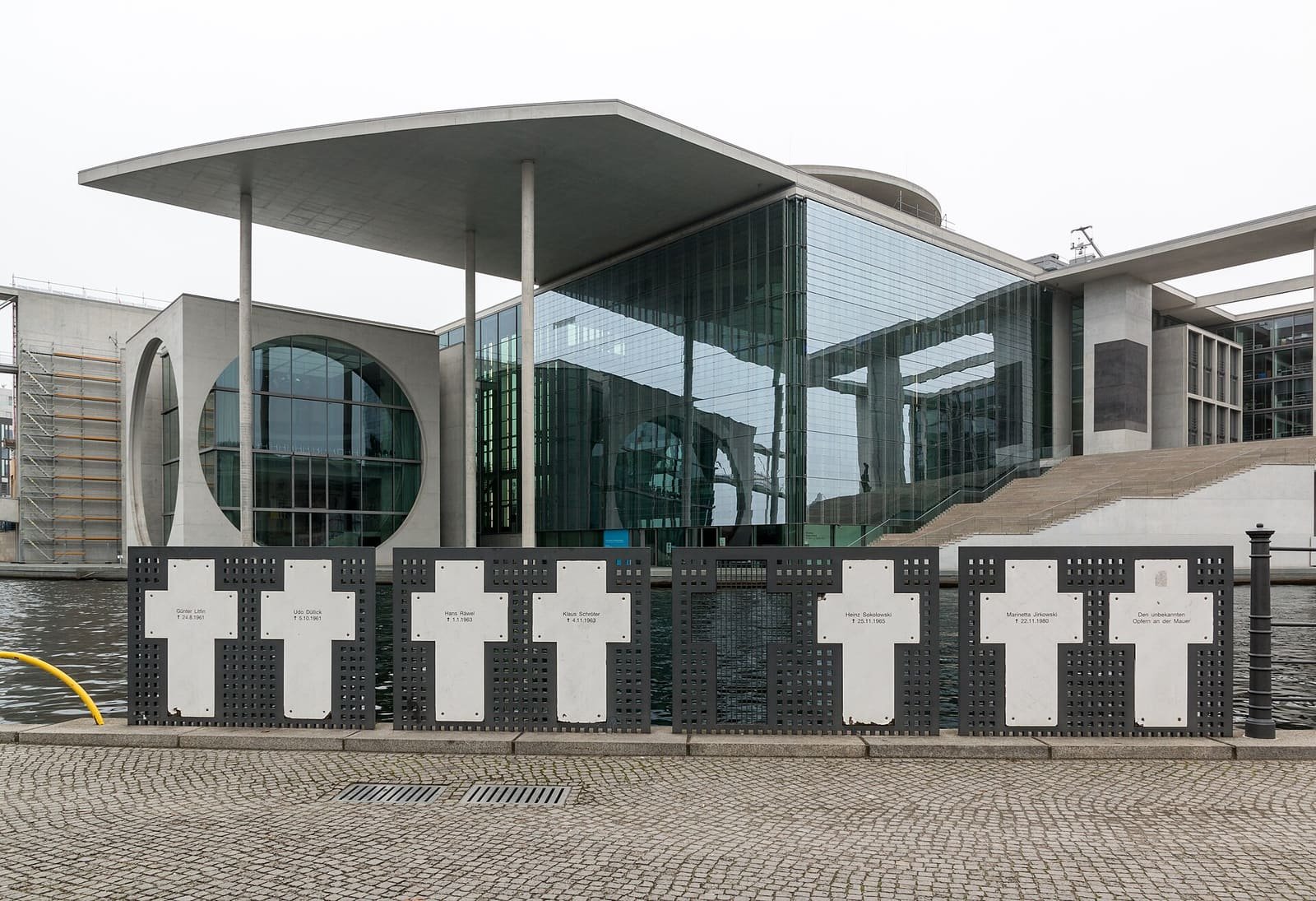 A row of white memorial crosses bearing names, on a riverside railing in front of a modern parliament building.