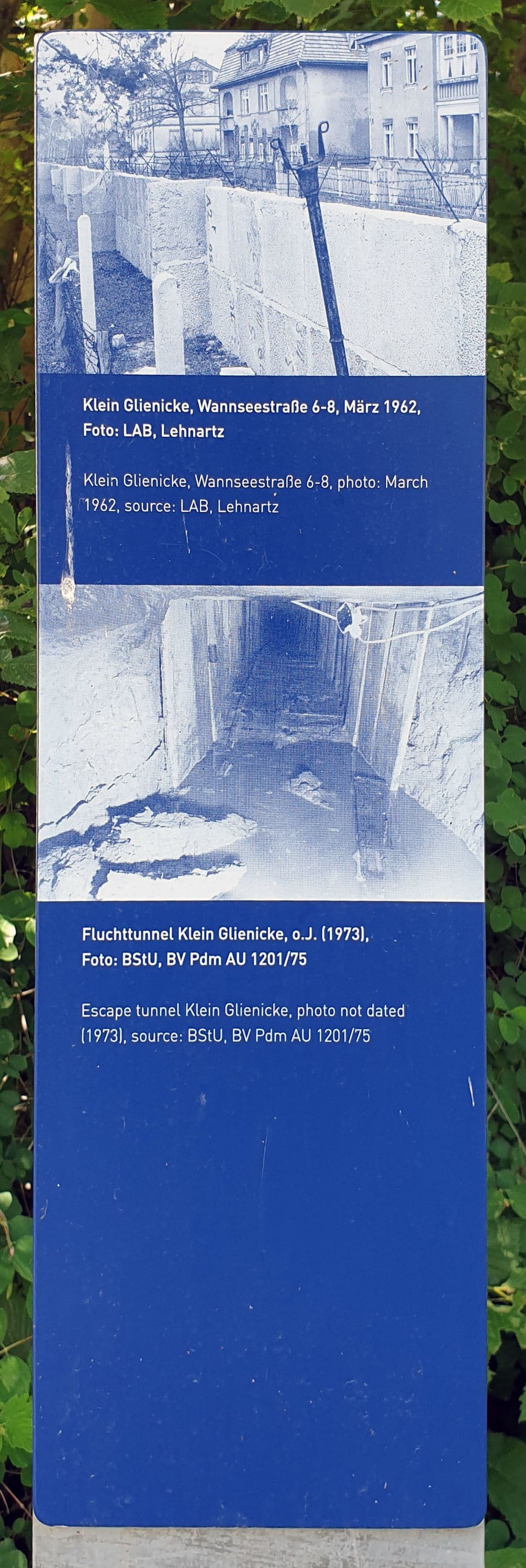 Tall blue information stele with two blue-tinted photos: the Wall beside villas, and a view into a narrow shored tunnel.