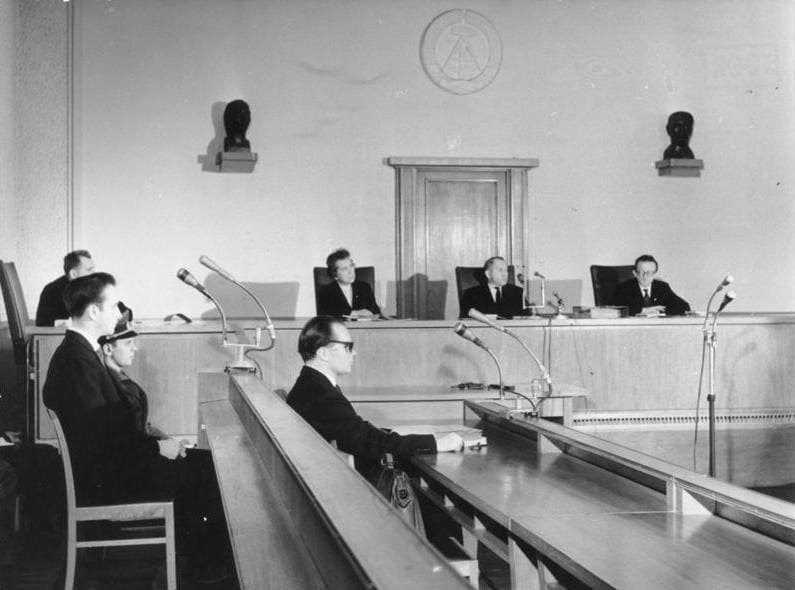 Black-and-white photo of a courtroom with judges seated at a raised bench, the defendant on the left and his lawyer in front.