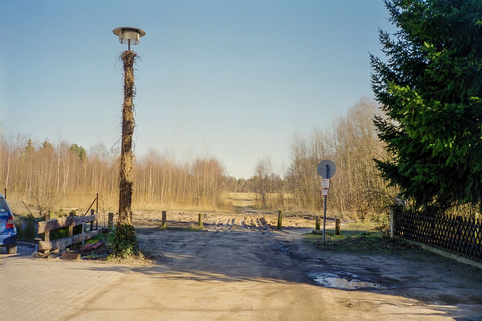A sandy road ending at open scrubland and trees, with a lamp post and a fence at the side.