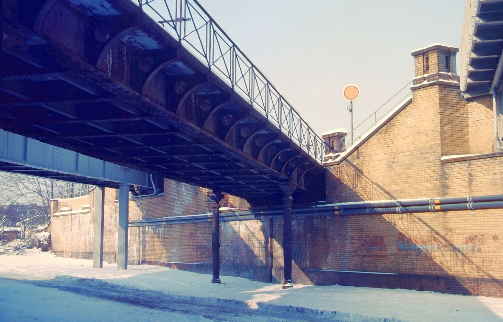 Colour photo of a snow-covered street passing under a steel railway bridge on iron columns, beside a high yellow-brick railway embankment with a round signal board on top.