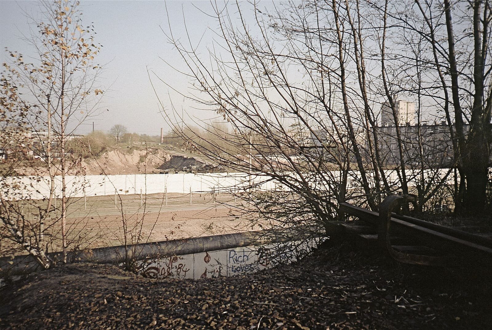 Colour photo through bare birch branches: a low wall in the foreground, a sandy border strip with a fence and a long white wall beyond, and an old railway buffer on the right.