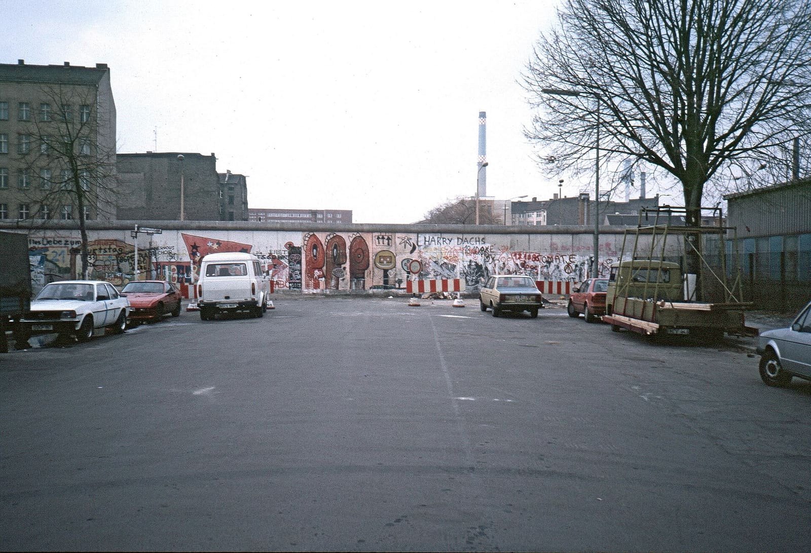 A wide cobbled street with parked cars running straight into a graffiti-covered Berlin Wall, with East Berlin buildings and a chimney behind it.