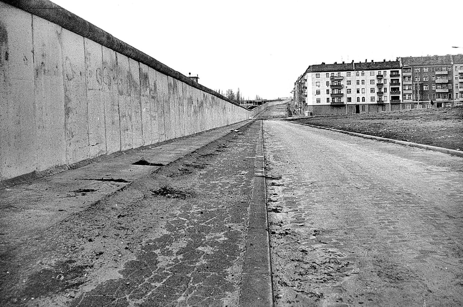 Black-and-white photo of a long, straight concrete wall on the left beside an empty cobbled road, with a row of apartment buildings on the right.