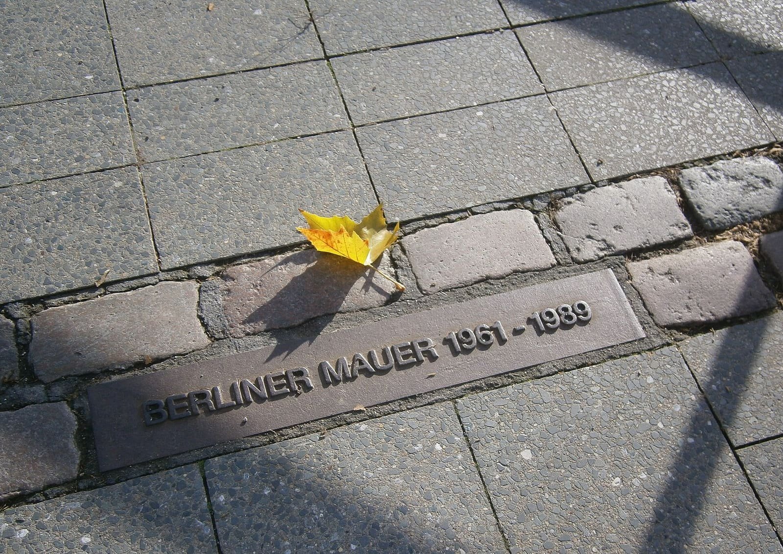 Colour photo of a row of cobbles in paving with a bronze strip reading 'Berliner Mauer 1961-1989' and a yellow autumn leaf.