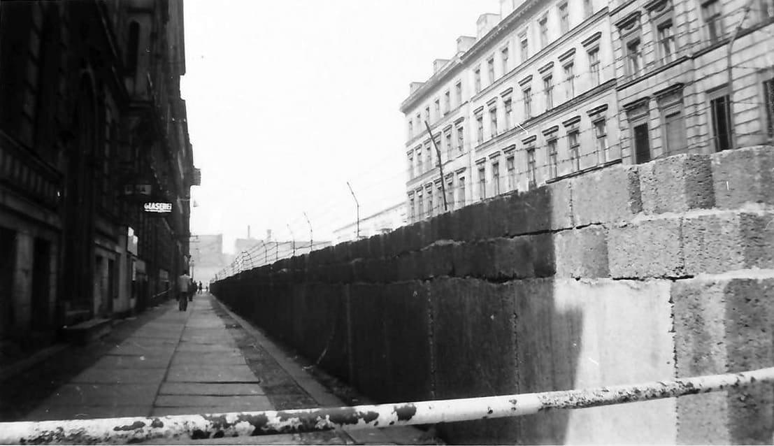 Black-and-white photo of a street with a wall running along it, apartment buildings on both sides.