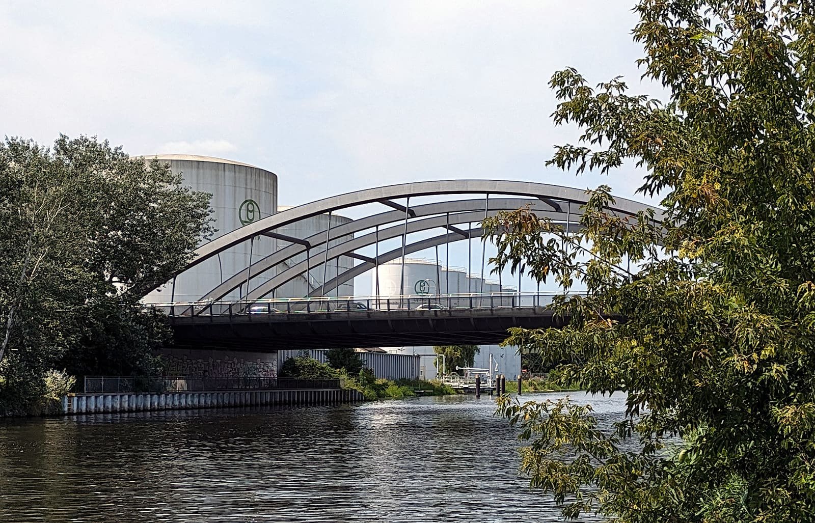 A modern steel arch bridge over a canal lined with trees, with large white storage tanks behind it.