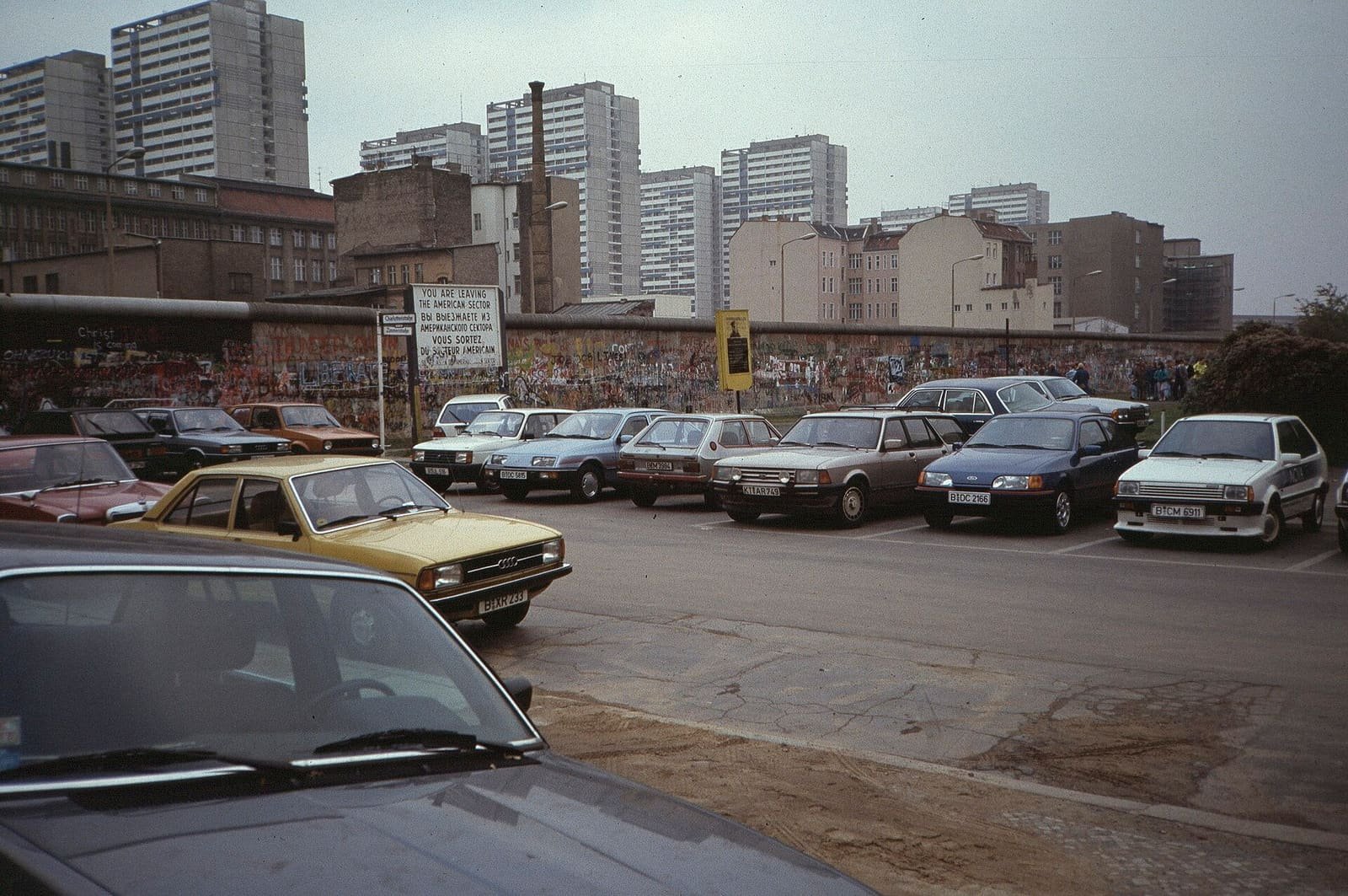 Colour photo: cars parked on a West Berlin lot in front of a graffiti-covered wall with a 'You are leaving the American sector' sign; behind the wall, old buildings and a row of tall white tower blocks.