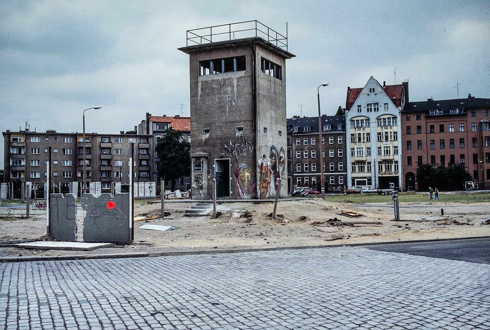 Colour photo: a square grey concrete watchtower with graffiti at its base stands on a sandy, cleared strip with a few leftover wall segments, in front of a row of apartment buildings under a cloudy sky.