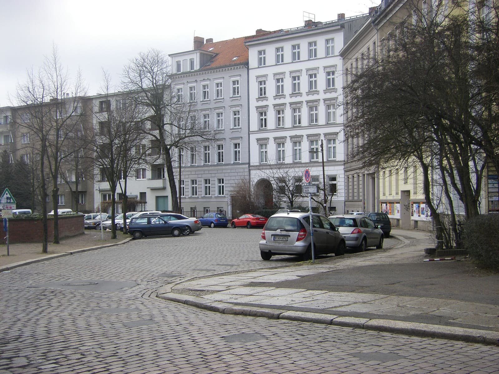 Colour photo: a cobbled street curves past parked cars towards old apartment buildings; a line of asphalt-filled holes crosses the cobbles in the foreground.