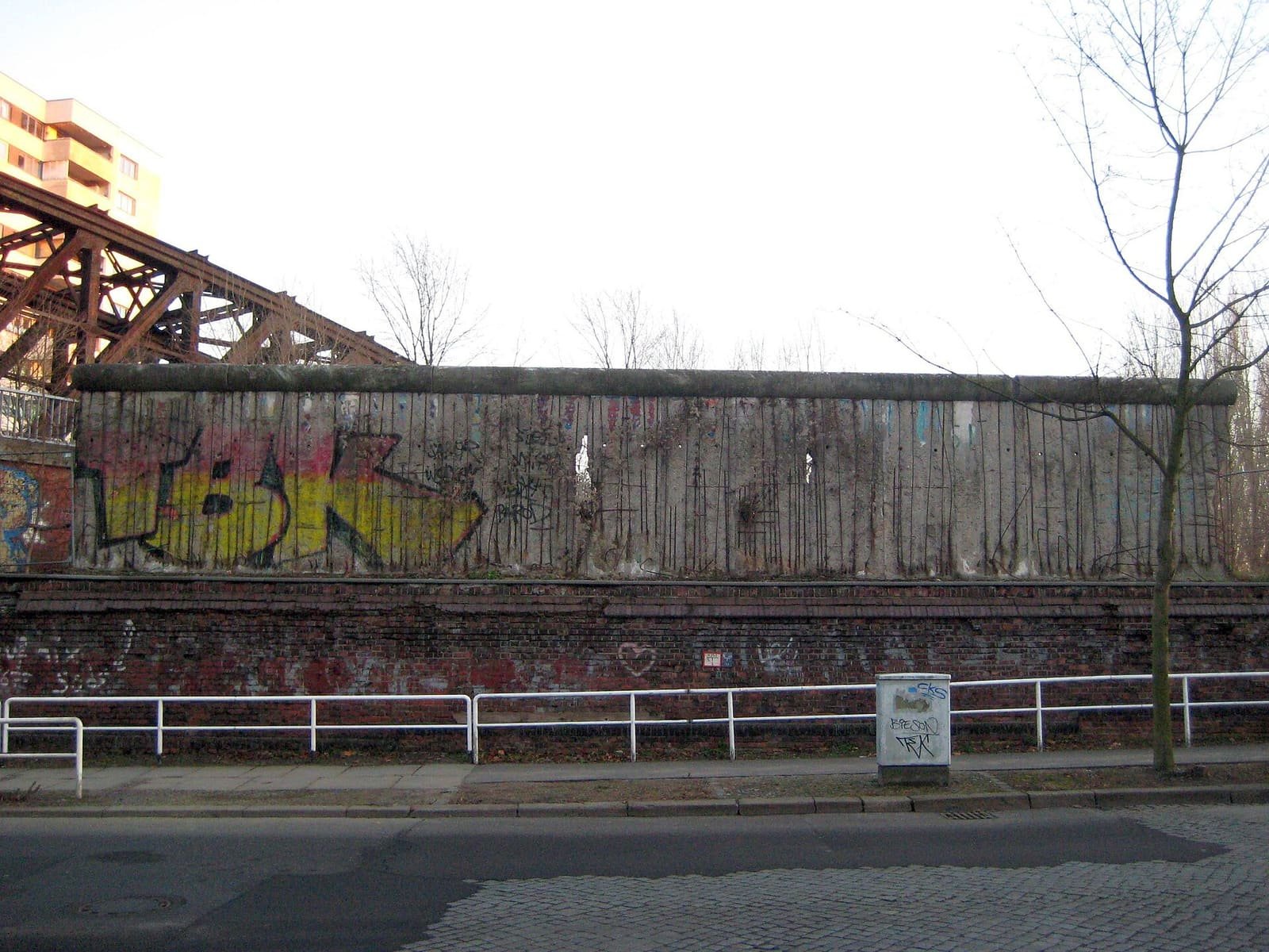 A long grey concrete wall streaked with graffiti, sitting on top of an older red-brick wall beside a street, with a steel railway bridge on the left.