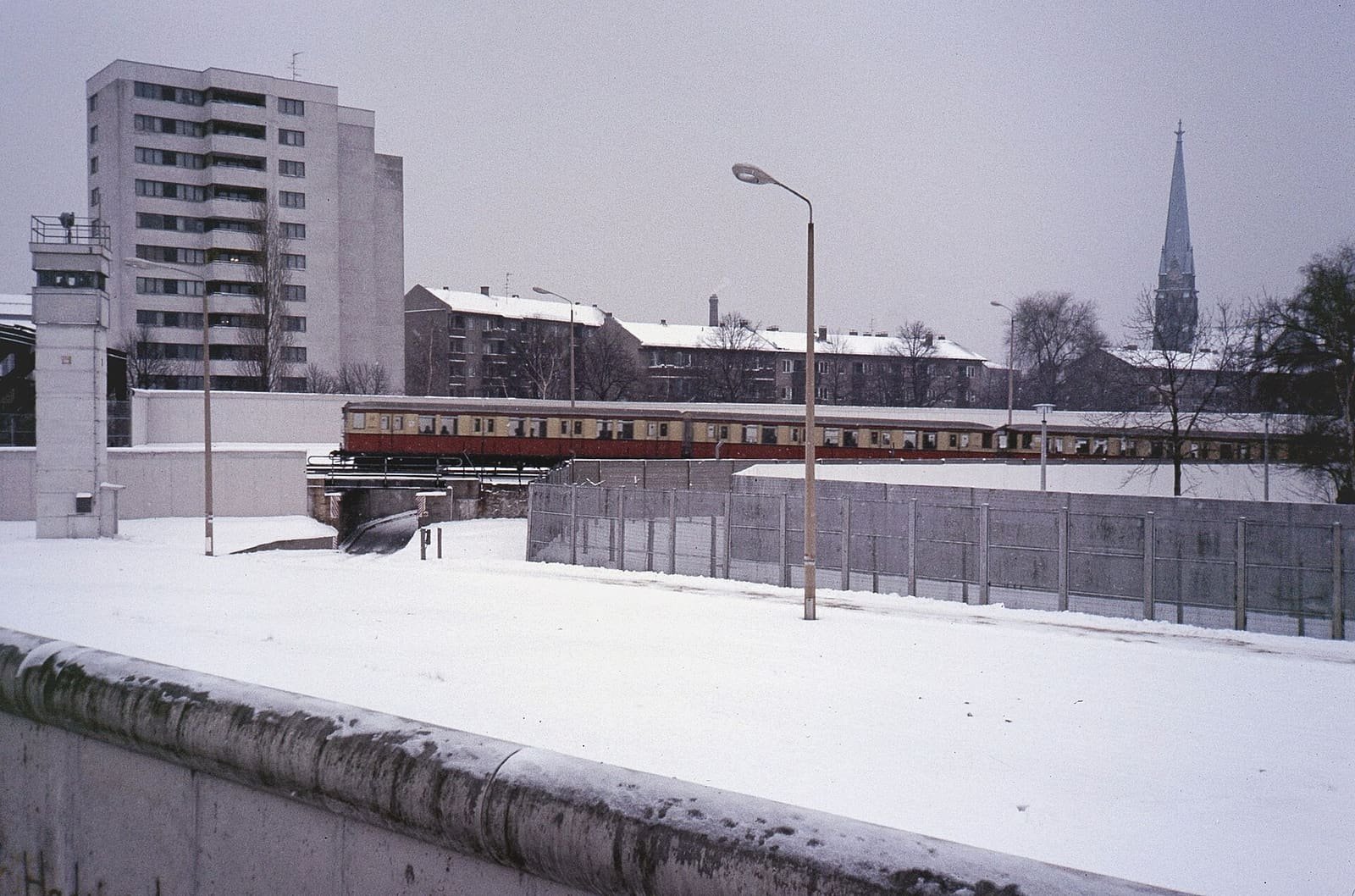 Snow-covered death strip seen over the top of the Wall, with a watchtower on the left, a red and cream S-Bahn train on a bridge and apartment blocks behind.