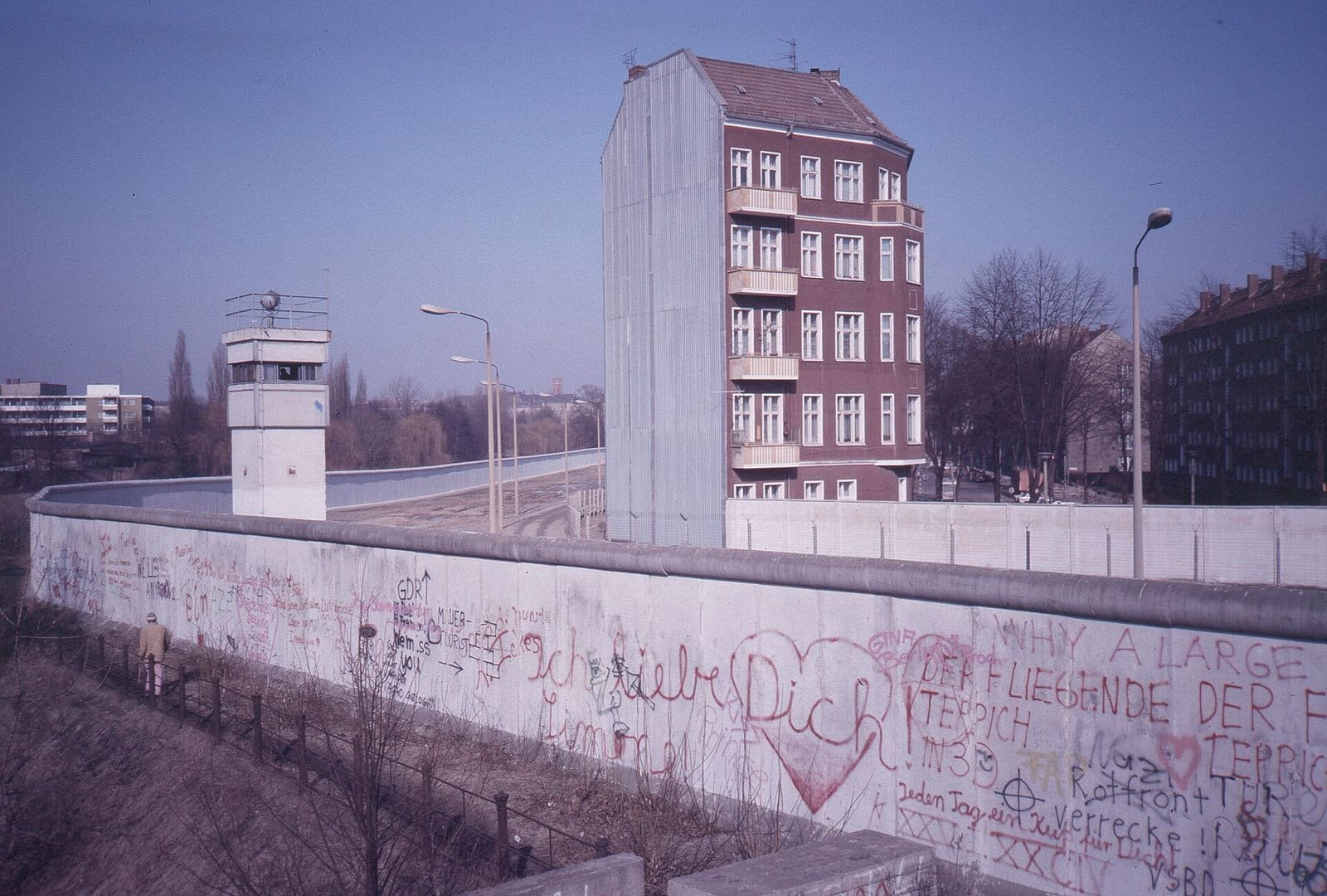 Colour photo from a raised bank over a long graffiti-covered wall: behind it a sandy border strip with a watchtower and lamp posts runs past a lone red-brick apartment house, with more buildings and trees beyond.