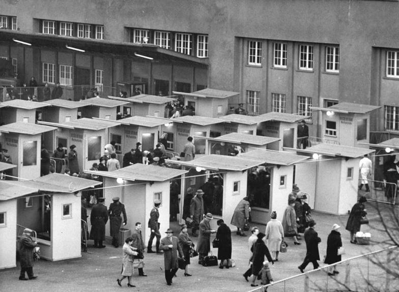 Black-and-white photo from above of rows of small white control booths with pitched roofs in a yard, with uniformed guards and travellers carrying bags.
