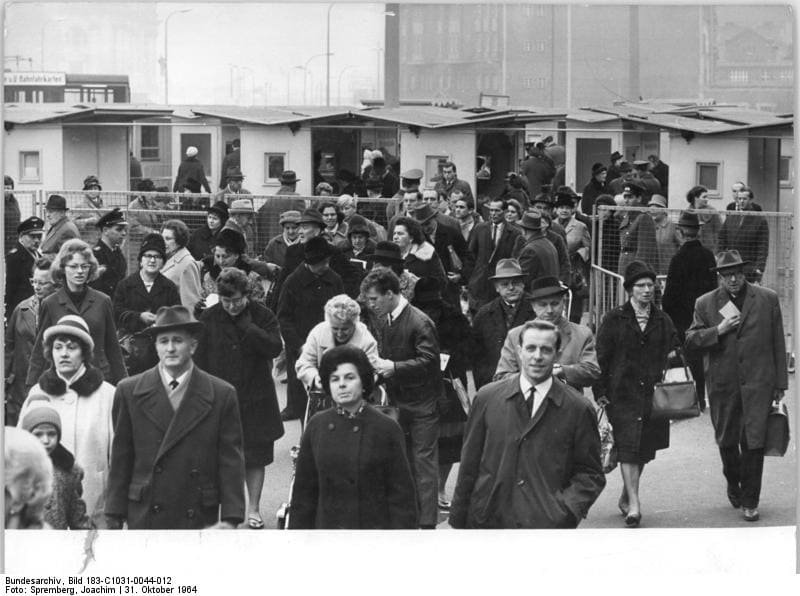 Black-and-white photo of a crowd of men and women in winter coats and hats walking towards the camera away from a row of small control booths.