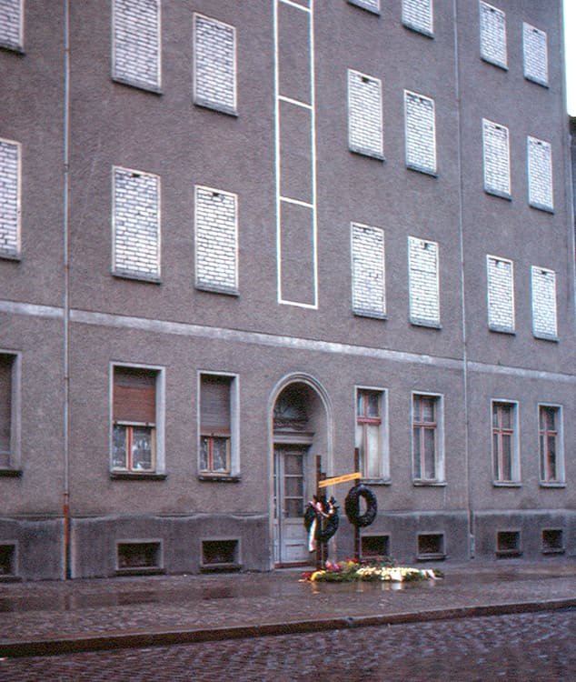 Historical photograph of a corner building with bricked-up windows and a small memorial with wreaths on the pavement