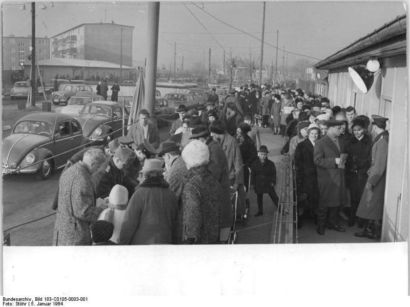 Black-and-white photo of a crowd in winter coats queuing between barriers beside a wooden hut, with parked cars and a uniformed guard.