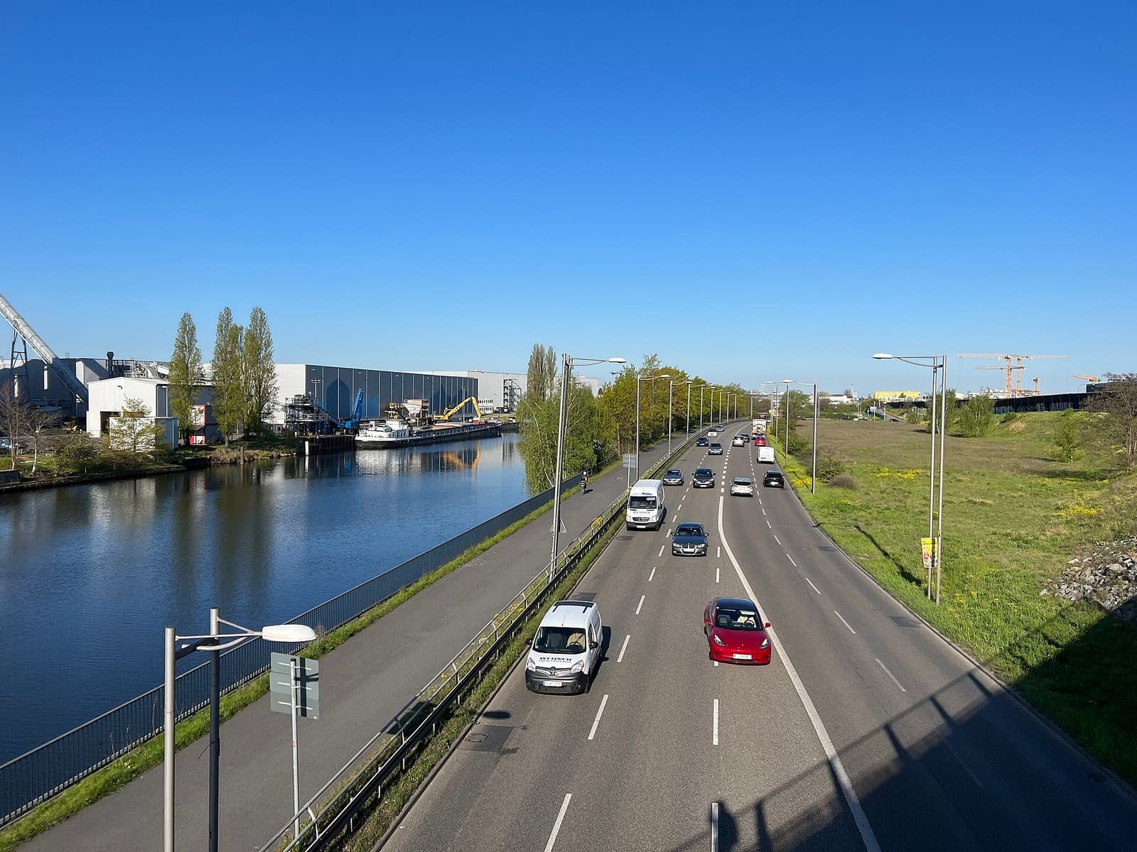 Colour photo from a bridge: a straight canal runs into the distance on the left, with industrial buildings and moored boats on its far bank; a multi-lane road with cars runs alongside it on the right under a clear blue sky.