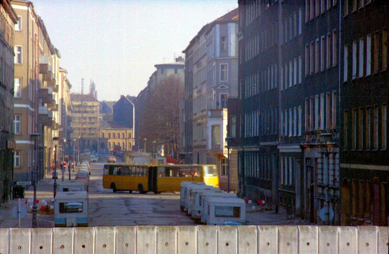 Colour photo taken over the top of a concrete wall: a cobbled street between tall apartment blocks, with a row of small white trailers parked in the border strip and a yellow articulated bus turning across the street further on.
