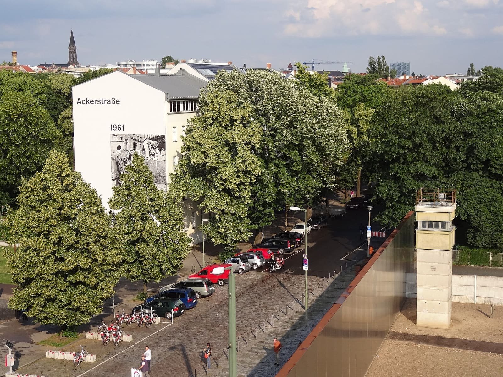 Colour photo from above: a cobbled street with parked cars and cyclists runs between leafy trees; on the right a rusty steel wall and a square concrete watchtower stand in a sandy strip, and a white gable wall with a large black-and-white photo rises on the left.